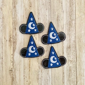 May include: Four blue wizard hat patches with white crescent moons and stars. The hats have black mouse ears and are outlined with white stitching. The patches are arranged on a light wood grain surface.