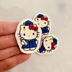 May include: Three embroidered patches featuring Hello Kitty in a graduation gown and cap. The patches have a white background with black outlines, a blue gown, and a red bow. One patch is held in a hand.