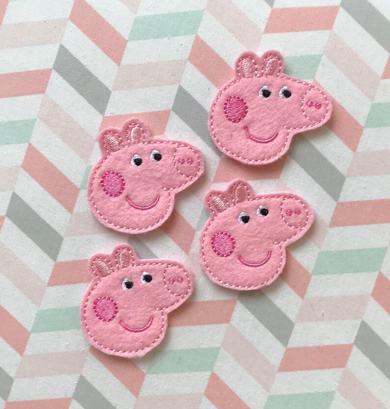 May include: Four pink felt embroidered patches shaped like the head of a cartoon pig. Each patch features two ears, two eyes, a snout with two nostrils, and a circular cheek detail. The embroidery is done in pink thread on a light pink felt base.