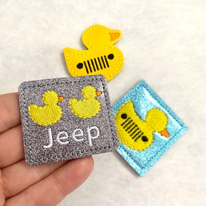 Peut inclure: Trois écussons brodés sur le thème du canard. Un écusson canard jaune avec un motif de calandre Jeep noire. Un écusson carré gris avec deux canards jaunes et le mot "Jeep". Un écusson losange bleu avec un canard jaune et une calandre Jeep.