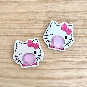 Peut inclure: Deux écussons blancs scintillants en forme de chat de dessin animé avec un nœud rose. Chaque écusson a un cercle rose irisé au centre et des moustaches noires. Les écussons sont sur une surface en bois brun clair.