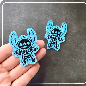 May include: Two light blue, glittery patches featuring a black skeleton design of a cartoon character. The patches are in the shape of the character, with a black outline. The patches are on a dark gray surface.