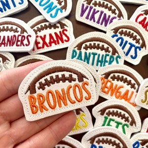 May include: A collection of embroidered patches shaped like footballs, each featuring a team name in various colors. The patches have a white border and brown football stitching. The word "BRONCOS" is visible in orange.