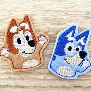 May include: Two embroidered felt patches featuring cartoon dog characters. One patch is tan and brown with white accents, while the other is light blue and dark blue with white details. Both patches depict stylized canine figures with raised arms.