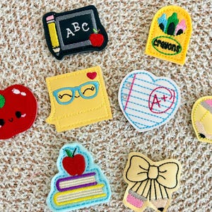 Peut inclure: Un ensemble de huit écussons brodés aux motifs scolaires comprenant un tableau noir avec ABC, une boîte de crayons jaune, une pomme rouge, un post-it jaune avec des lunettes, un papier en forme de cœur avec A+, un crayon, des livres et un nœud.