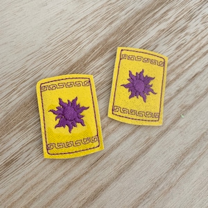 May include: Two yellow rectangular patches with a purple sunburst design. Each patch has a decorative border and a textured appearance. The patches are placed on a light-colored wooden surface.