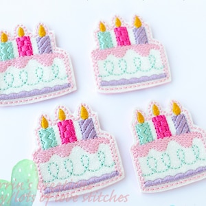 May include: Four embroidered birthday cake patches featuring white frosting, pink icing drips, and three candles in mint, pink, and purple with yellow flames. Each patch has a scalloped purple base and decorative mint green swirl stitching.