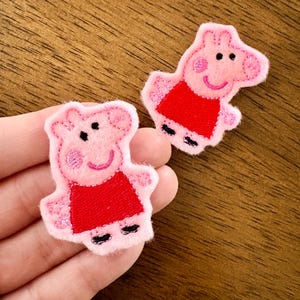 May include: Two embroidered fabric patches shaped like a cartoon pig character. Each patch features a light pink pig with a red dress, black outlines for facial features, and small black feet. The embroidery has a textured finish on the surface.