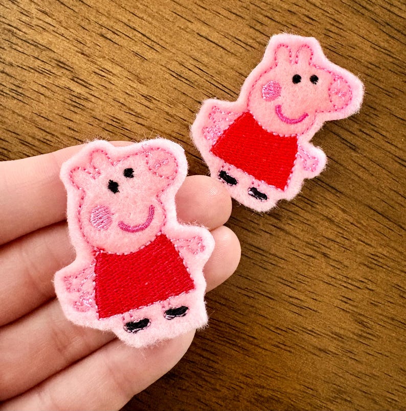 May include: Two embroidered fabric patches shaped like a cartoon pig character. Each patch features a light pink pig with a red dress, black outlines for facial features, and small black feet. The embroidery has a textured finish on the surface.