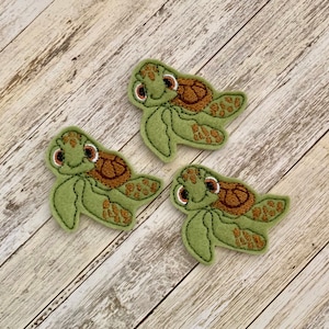 May include: Three green turtle-shaped felt appliques with brown shell details and embroidered features. Each turtle has a smiling face with large eyes. The appliques are arranged on a light wood-grain surface.
