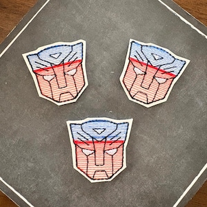 May include: Three embroidered patches featuring the Autobot insignia from the Transformers franchise. Each patch is shaped like a stylized robot face with a blue upper section, a red lower section, and white accents, all outlined in black thread.
