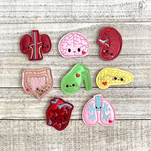 May include: A collection of seven embroidered patches featuring stylized human organs including a dark red kidney pair, a pink brain, a dark red kidney, a tan intestine, a green gallbladder, a yellow pancreas, a dark red heart, and pink lungs.