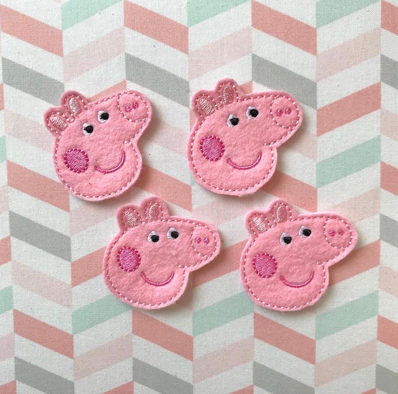 May include: Four pink felt fabric appliques shaped like the head of a cartoon pig. Each piece features embroidered details including two eyes, a snout with two nostrils, a smiling mouth, two ears, and a circular cheek patch in a darker pink thread.