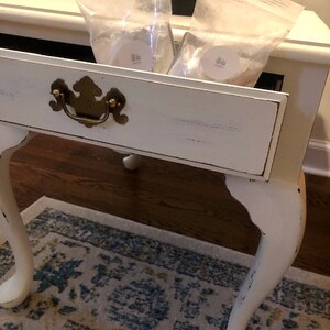 May include: A white end table with a single drawer and turned legs. The drawer is open and contains two clear plastic bags with white labels. The table is sitting on a patterned rug.