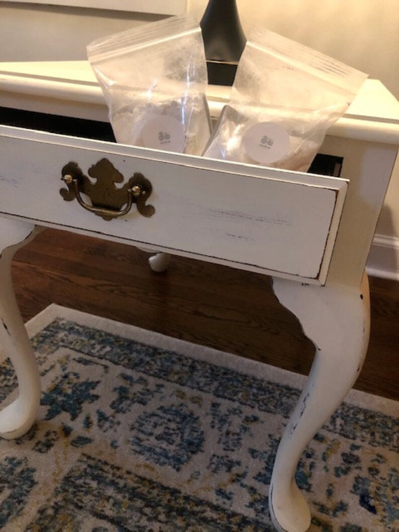 May include: A white end table with a single drawer and turned legs. The drawer is open and contains two clear plastic bags with white labels. The table is sitting on a patterned rug.