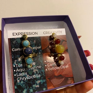 May include: Two gold wire wrapped rings with different colored gemstones. The ring on the left has turquoise, aquamarine, lapis lazuli, and chrysocolla stones. The ring on the right has white, red, and yellow stones. The rings are in a purple box with a white card that says "Expression" and "Creativity" with a list of the stones used.