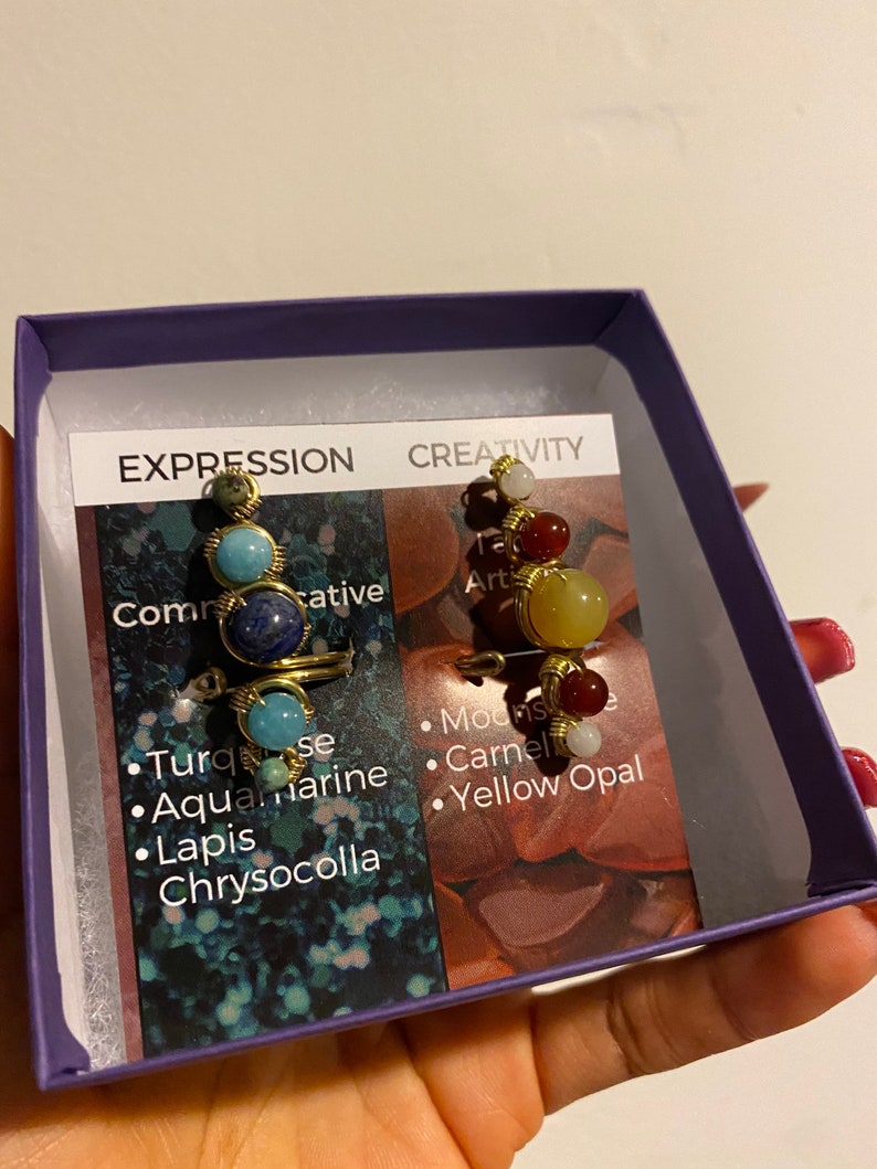 May include: Two gold wire wrapped rings with different colored gemstones. The ring on the left has turquoise, aquamarine, lapis lazuli, and chrysocolla stones. The ring on the right has white, red, and yellow stones. The rings are in a purple box with a white card that says "Expression" and "Creativity" with a list of the stones used.