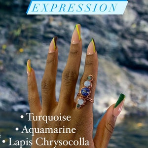 May include: A hand with long, light-colored nails with green and yellow accents. The hand is wearing a ring with turquoise, aquamarine, and lapis chrysocolla stones. The text "EXPRESSION" is at the top of the image. The text "Turquoise", "Aquamarine", and "Lapis Chrysocolla" is at the bottom of the image.
