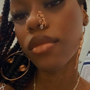 May include: A person wearing a gold chain necklace with a white and red stone pendant. They are also wearing a gold nose ring and earrings. The person has dark skin and is wearing their hair in braids.