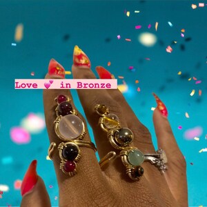 May include: A close-up of a hand wearing four gold rings with different colored gemstones. The rings are made with wire wrapped around the stones. The text "Love in Bronze" is visible in the image.