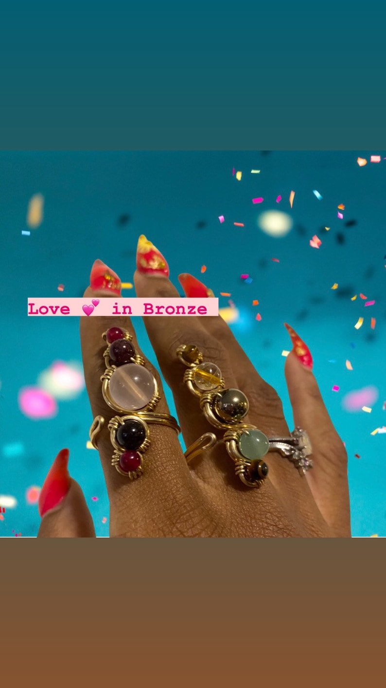 May include: A close-up of a hand wearing four gold rings with different colored gemstones. The rings are made with wire wrapped around the stones. The text "Love in Bronze" is visible in the image.