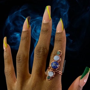 May include: A hand with long, almond-shaped nails painted with a green and yellow design. The hand is wearing a ring with multiple stones, including blue, green, and white.
