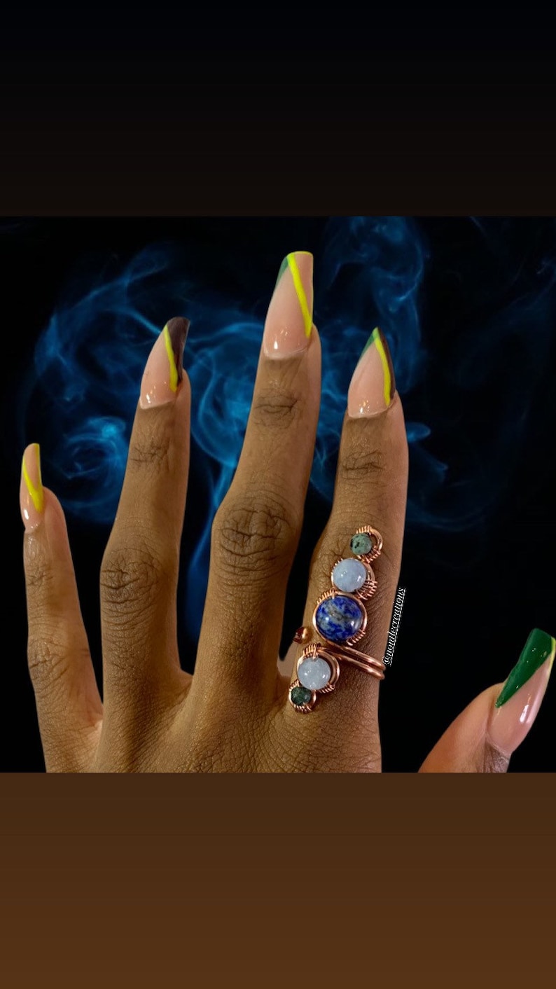 May include: A hand with long, almond-shaped nails painted with a green and yellow design. The hand is wearing a ring with multiple stones, including blue, green, and white.