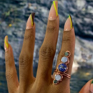 May include: A close-up of a hand with long, almond-shaped nails painted with a brown and yellow design. The hand is wearing a ring with three stones: a blue lapis lazuli, a white moonstone, and a green aventurine.