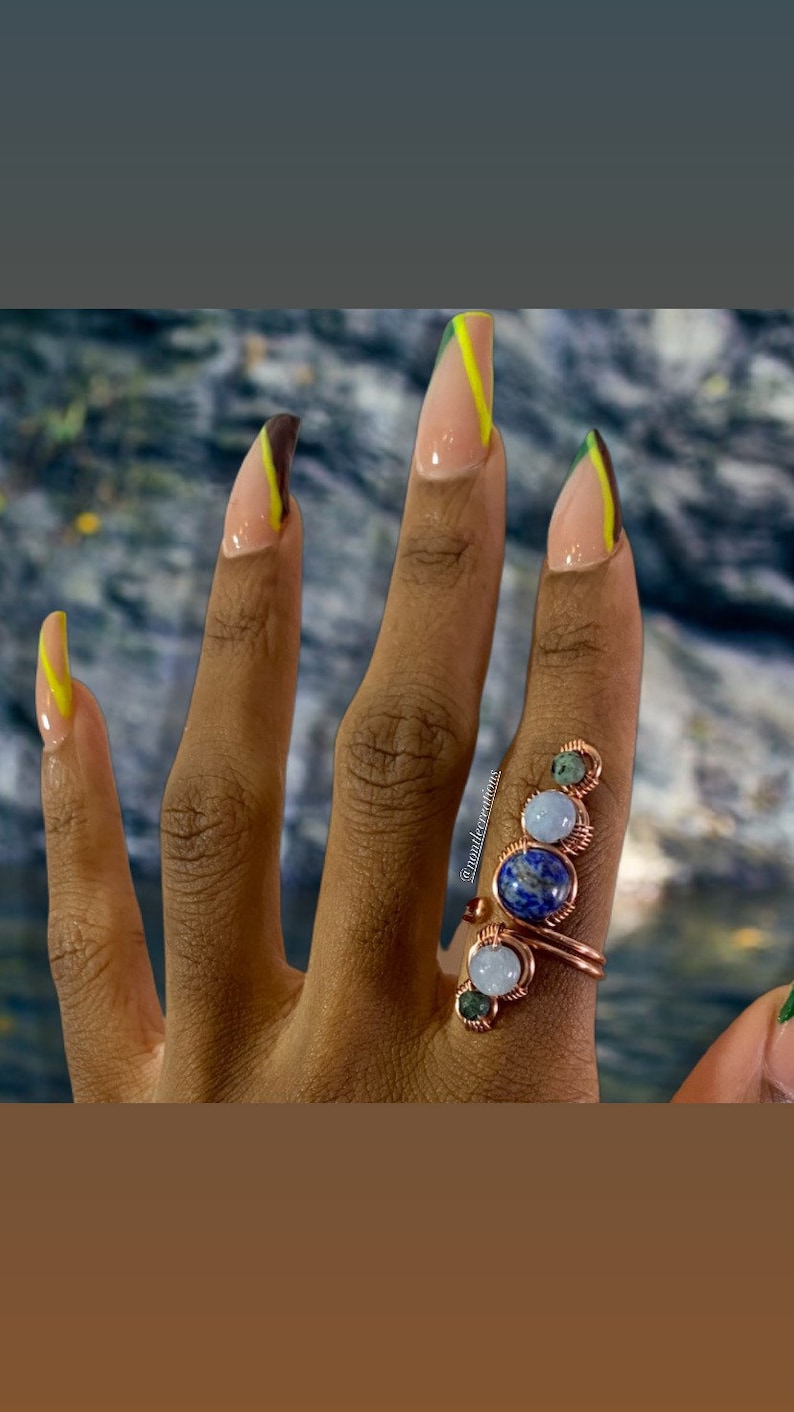 May include: A close-up of a hand with long, almond-shaped nails painted with a brown and yellow design. The hand is wearing a ring with three stones: a blue lapis lazuli, a white moonstone, and a green aventurine.