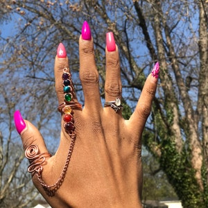 May include: A hand wearing a copper ring with a chain and seven colorful gemstones. The ring is on the index finger. The nails are painted pink and have a pointed shape.