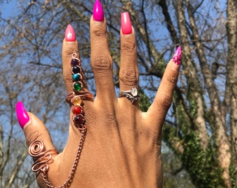Puede incluir: Una mano con un anillo de cobre con una cadena y siete piedras preciosas de colores. El anillo está en el dedo índice. Las uñas están pintadas de rosa y tienen una forma puntiaguda.