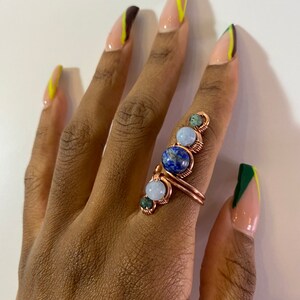 May include: A copper wire ring with four stones: a blue lapis lazuli, a light blue aquamarine, and two green stones. The ring is on a hand with light brown skin and nails painted with a green and yellow design.