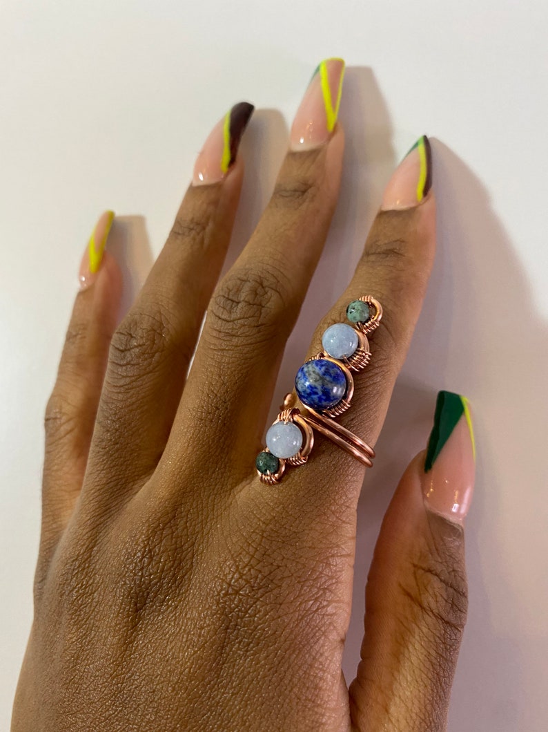 May include: A copper wire ring with four stones: a blue lapis lazuli, a light blue aquamarine, and two green stones. The ring is on a hand with light brown skin and nails painted with a green and yellow design.