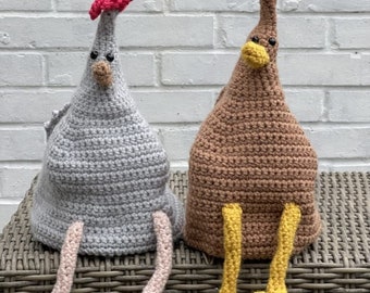 May include: Two crocheted chicken figurines, one gray with pink legs and one brown with yellow legs, sitting on a wicker surface.
