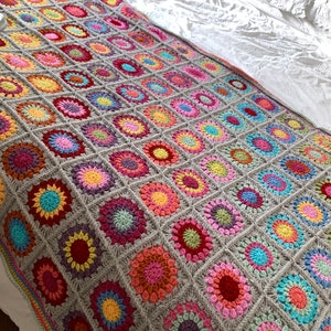 May include: A colourful crocheted throw blanket with a grey background and a variety of bright colours, including pink, red, yellow, blue, and green. The blanket is made up of a series of squares, each with a different pattern.