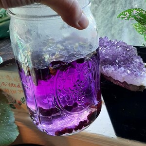 May include: A clear glass mason jar filled with vibrant purple liquid and small, dark red objects. The jar is held by a hand and is next to a large, purple amethyst crystal cluster in a black pot.