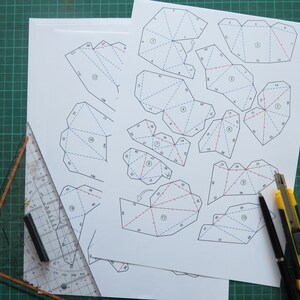 May include: A printed paper template with numbered lines and red and blue markings. The template is for a 3D paper model. A ruler, a pen, and a utility knife are on a green cutting mat.