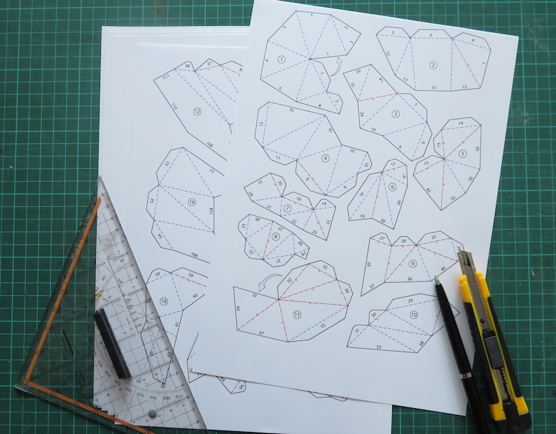 May include: A printed paper template with numbered lines and red and blue markings. The template is for a 3D paper model. A ruler, a pen, and a utility knife are on a green cutting mat.