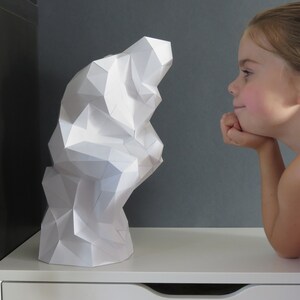 May include: A white paper sculpture of a geometric shape, resembling a cloud or a mountain, is viewed by a young girl with a curious expression.