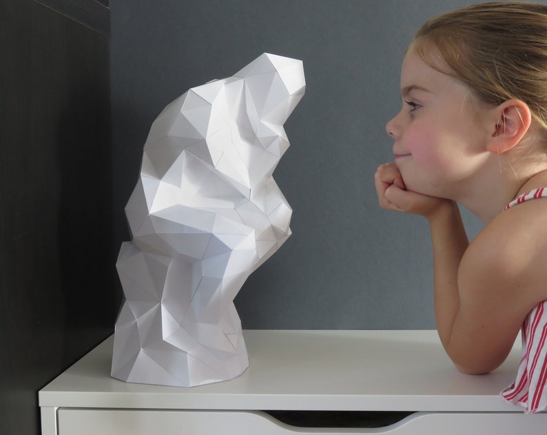 May include: A white paper sculpture of a geometric shape, resembling a cloud or a mountain, is viewed by a young girl with a curious expression.