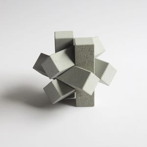 Puede incluir: Escultura geométrica compuesta por bloques rectangulares entrelazados. Los bloques son principalmente de color gris claro, algunos con una superficie gris más oscura y texturizada. La disposición crea una forma abstracta tridimensional sobre un fondo blanco.