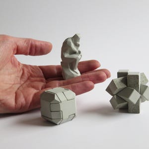 Op de afbeelding: Drie geometrische betonnen sculpturen in grijstinten. Eén is een kubus, een andere een figuur en de derde een cluster van blokken. De sculpturen worden op een wit oppervlak getoond.