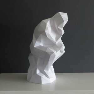 May include: A white paper sculpture of a person sitting with their chin on their hand, in a thinking pose. The sculpture is made of many small triangles and is a modern interpretation of a classic sculpture.