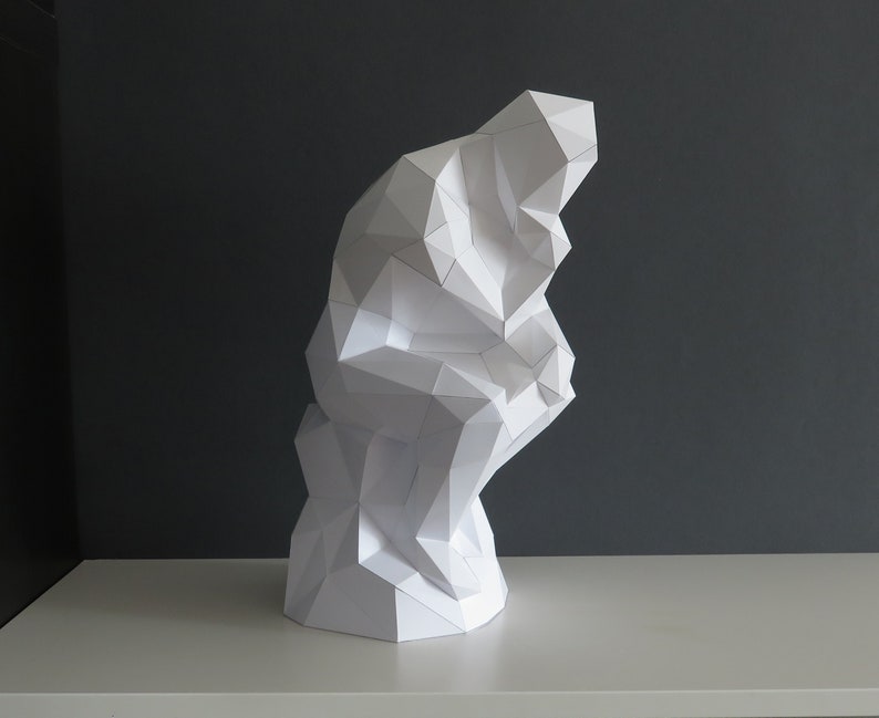 May include: A white paper sculpture of a person sitting with their chin on their hand, in a thinking pose. The sculpture is made of many small triangles and is a modern interpretation of a classic sculpture.