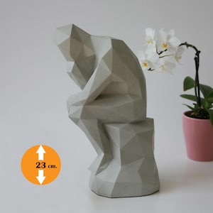 Könnte beinhalten: Graue Betonskulptur einer sitzenden, nachdenklichen Person. Die Statue ist 23 cm hoch und hat ein geometrisches, facettiertes Design. Eine weiße Orchidee in einem rosa Topf befindet sich im Hintergrund.