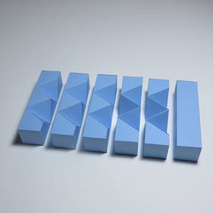 May include: A set of seven light blue geometric puzzle pieces made of paper. The pieces are arranged in a row and are all the same size and shape.