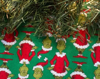 May include: Green Christmas tree skirt with a repeating pattern of the Grinch wearing a Santa hat. The skirt is made of fabric and has a white trim.