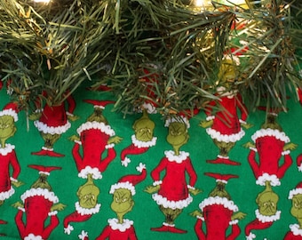 May include: A green Christmas tree skirt with a repeating pattern of the Grinch wearing a Santa hat. The skirt is made of fabric and has a white trim.