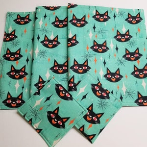 May include: A set of aqua-colored kitchen linens featuring a repeating pattern of black cat faces with orange eyes and smiles, and starburst and diamond shapes. The set includes placemats and square coasters.