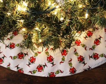 May include: White Christmas tree skirt with a repeating pattern of the Grinch wearing a Santa hat. The skirt is decorated with a green and red color scheme.